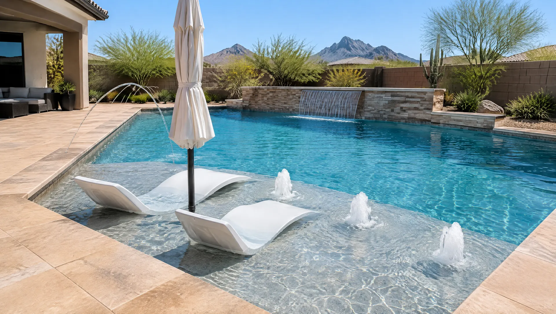 New pool with a large baja shelf holding an umbrella sleeve and two in-water loungers, three bubblers on the shelf, two deck jets arcing into the pool, sheer descent from a low wall, warm sandstone deck, clear Arizona sky