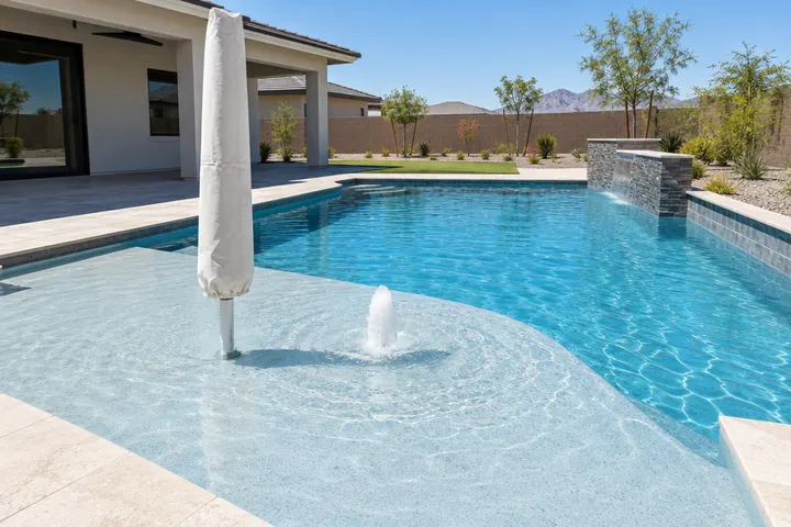 Large shallow tanning ledge at one end of a new pool, a few inches of water over light blue finish, umbrella sleeve and bubbler visible