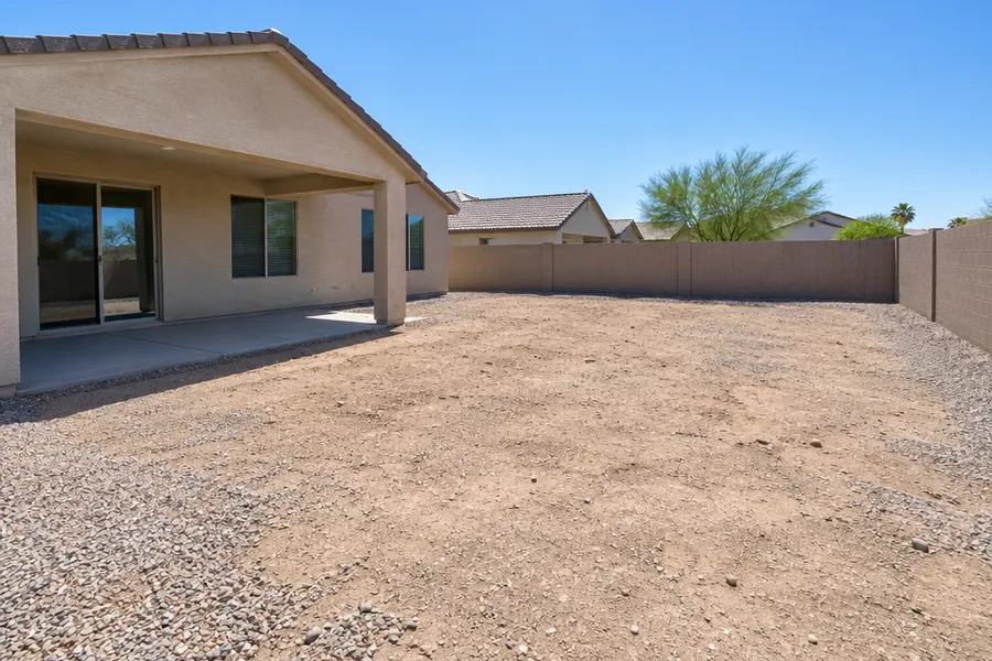 Empty Mesa, Arizona backyard before a pool is built: bare dirt and patchy gravel, a small plain concrete patio under a covered patio, block wall, single-story stucco home, harsh midday sun, no pool