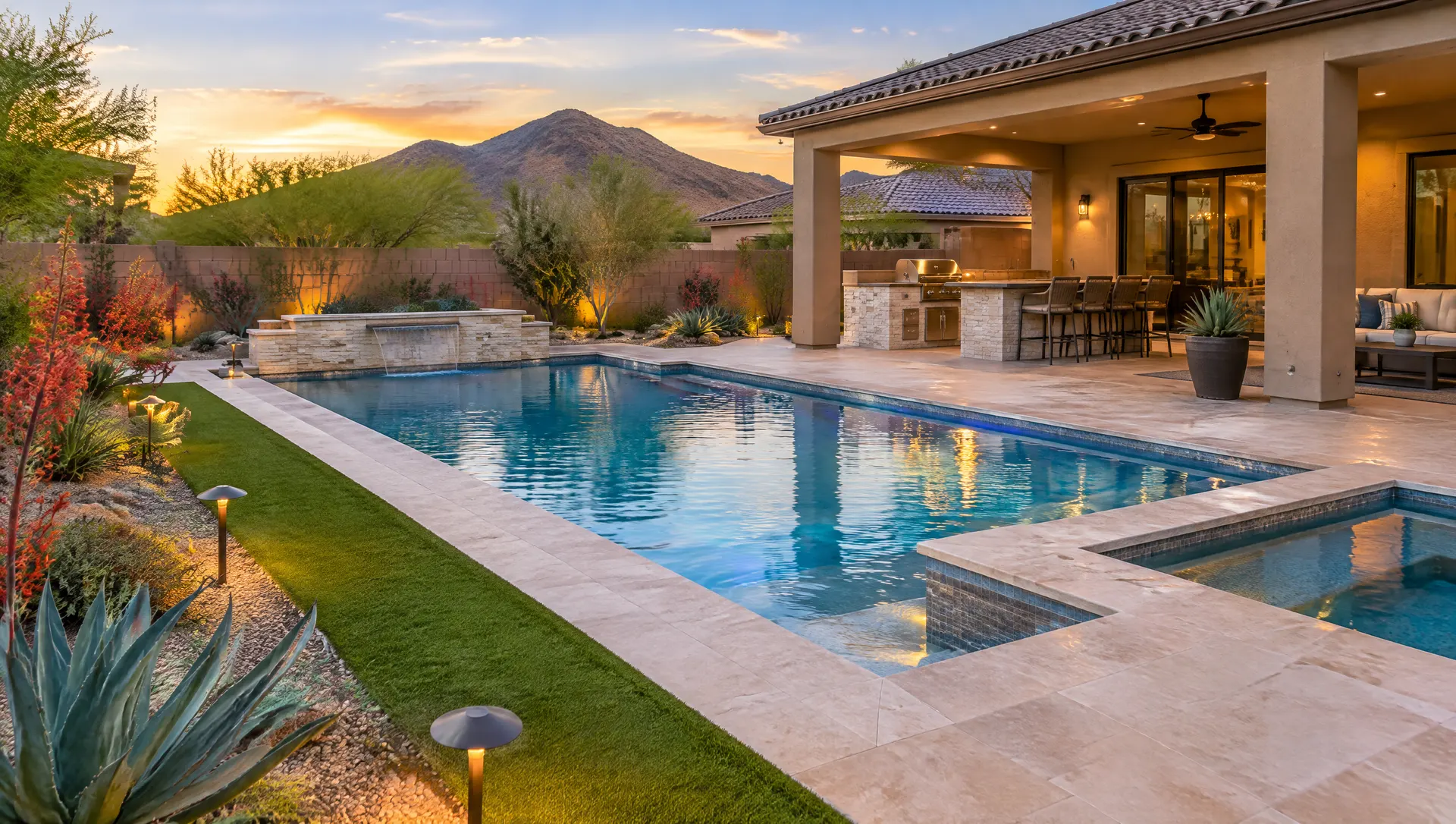 Finished new backyard around a brand-new pool in Mesa: light sandstone-color cool deck and travertine coping, artificial turf strip, desert plantings with agave and red yucca, a built-in barbecue island under a covered patio, low landscape lights, evening glow